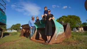 A family - two adults and two children, standing outside in front of machinery on a sunny day.