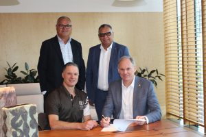 Four men, two are standing and two are sitting at a desk with one signing a sheet of paper.